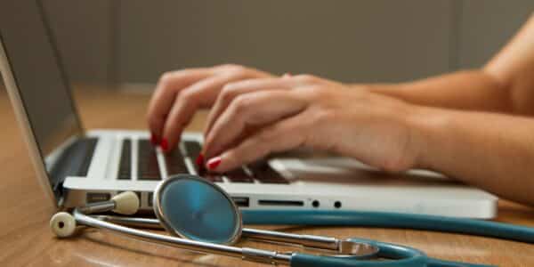 Person typing on a laptop with a stethoscope near the laptop.